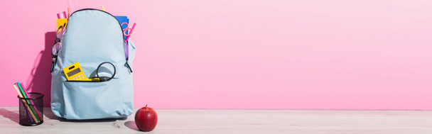 horizontales Bild eines blauen Rucksacks mit Schulbedarf in der Nähe des ganzen Apfels und Stifthalter mit Filzstiften auf rosa - Foto, Bild