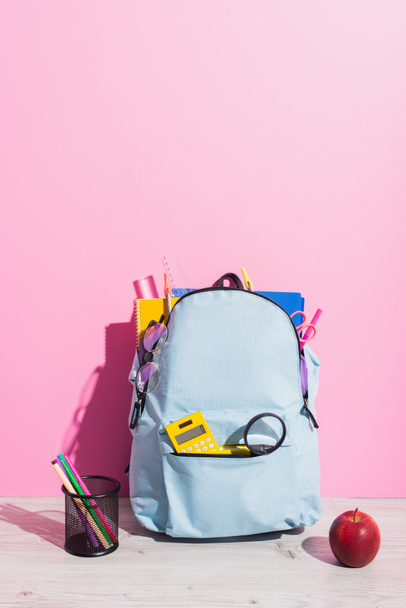 sac à dos bleu rempli de fournitures scolaires près de pomme mûre et porte-stylo avec stylos en feutre sur rose - Photo, image