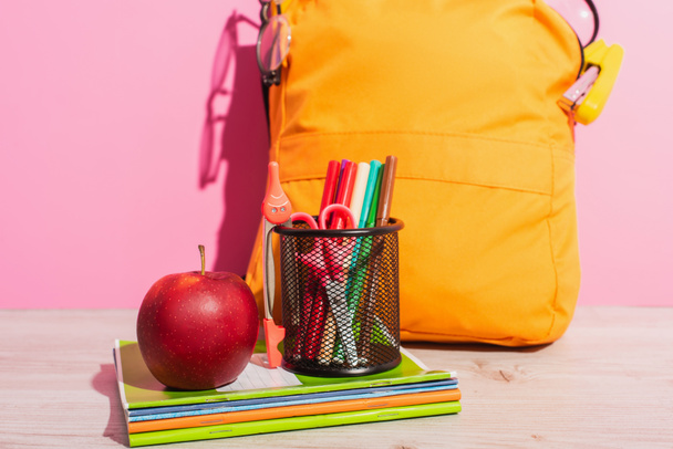 packed school backpack near notebooks, pen holder with felt pens, scissors and ripe apple on pink - Photo, Image