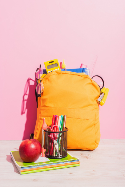 packed school backpack near notebooks, pen holder with felt pens and scissors, and ripe apple on pink - Fotografie, Obrázek