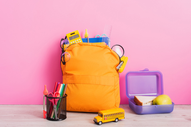 gele rugzak met schoolbriefpapier in de buurt van speelgoed schoolbus, lunchbox en pennenhouder met vilten pennen en schaar op roze - Foto, afbeelding