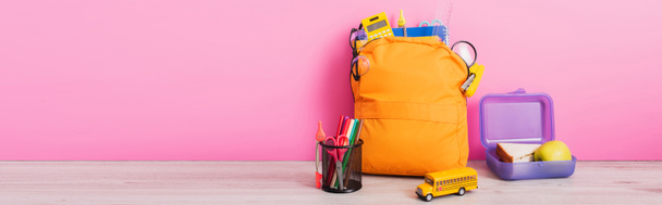 horizontal image of yellow backpack with school stationery near lunch box, toy school bus and pen holder with felt pens on pink - Photo, Image