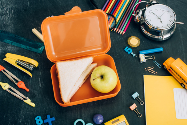 bovenaanzicht van lunchbox met rijpe appel en broodjes in de buurt van vintage wekker en schoolbriefpapier op zwart schoolbord - Foto, afbeelding
