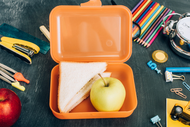 вид на обеденную коробку с бутербродами и спелое яблоко возле школьных канцелярских принадлежностей на черной доске - Фото, изображение