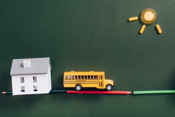 top view of yellow school bus on road made of color pencils, house model and sun made of magnets on green chalkboard - Фото, изображение