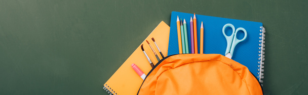 horizontal image of notebooks, color pencils, scissors and paintbrushes in yellow backpack on green chalkboard - Photo, Image