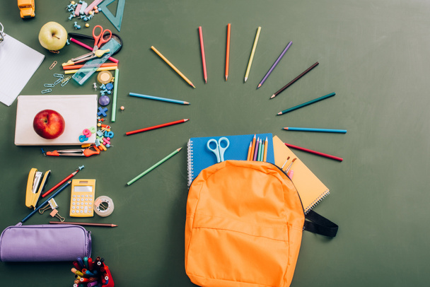 bovenaanzicht van rugzak met notitieboekjes, kleurpotloden en schaar in de buurt van schoolbenodigdheden op groen schoolbord - Foto, afbeelding