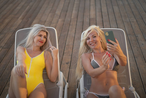 Piękne młode kobiety w bikini relaksujące się na leżakach z koktajlami i robiące selfie. Lato, zabawa, pomysł na wakacje. przestrzeń do kopiowania, miejsce na tekst - Zdjęcie, obraz
