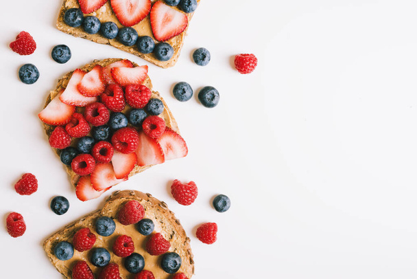 Breakfast toasts with wholegrain bread, peanut butter, strawberry, blueberry and raspberry on white table, top view. Healthy breakfast - Photo, Image