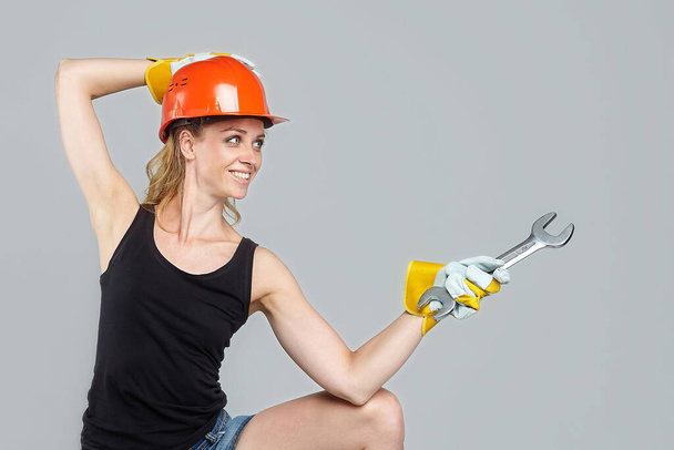 blonde woman in a protective helmet and gloves, holding a large wrench in his hands, points to an empty space. - Foto, afbeelding