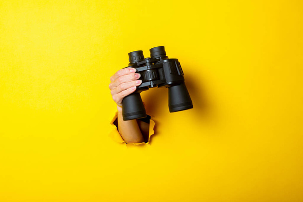 Mão feminina segura binóculos pretos no fundo amarelo, viajar, encontrar e conceito de pesquisa
. - Foto, Imagem