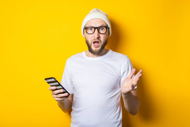 Bearded young man in a hat and glasses holding a phone in surprise - Foto, imagen