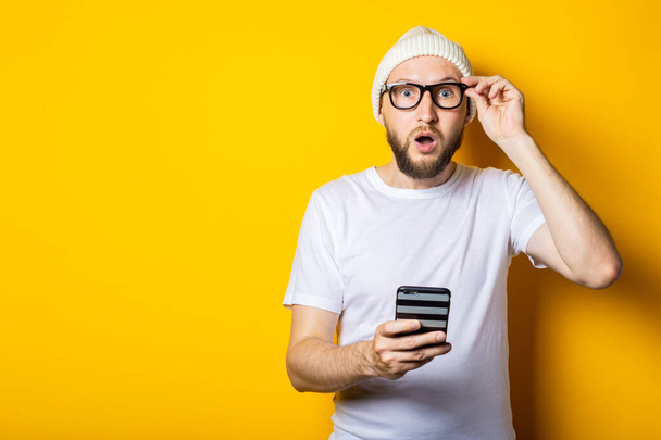 Bearded young man in shock surprised looks at the phone on a yellow background. - Fotografie, Obrázek