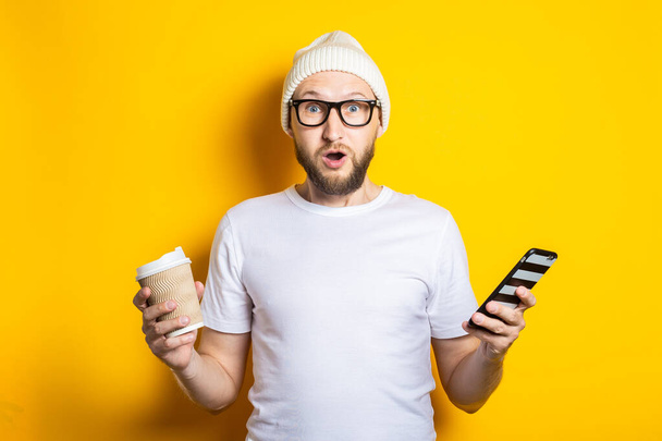 Překvapený vousatý mladý muž s lepenkovým sklem a kávou, drží telefon na žlutém pozadí - Fotografie, Obrázek