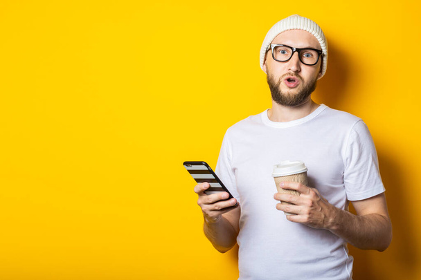 Surprised shocked bearded young man holding a phone and a cardboard glass with coffee on a yellow background. - Photo, image