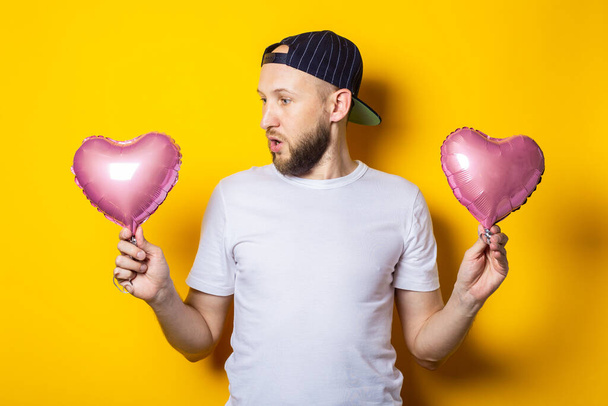 Бородатый молодой человек держит розовые воздушные шары сердца на желтом фоне - Фото, изображение
