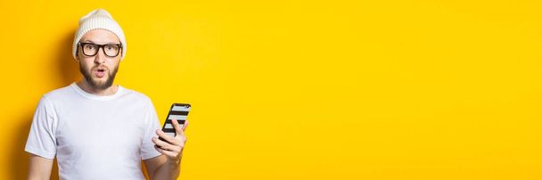 Surprised shocked young man with a beard holding a phone on a yellow background. Banner - Photo, image