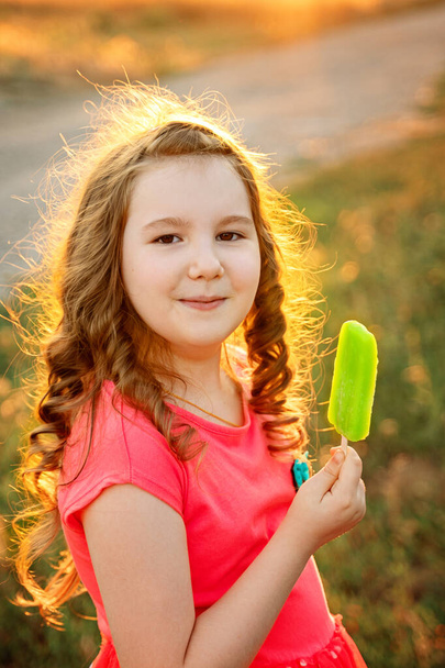 Linda niña rizada sonriente en vestido rosa con helado verde en su brazo hacia fuera en la puesta de sol. Actividades con niños verano al aire libre. Vertical
. - Foto, Imagen