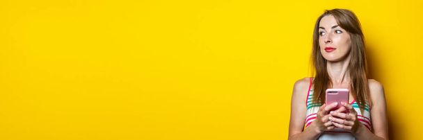 Pensive cute young woman with the phone looks up on a yellow background. Banner - Photo, Image
