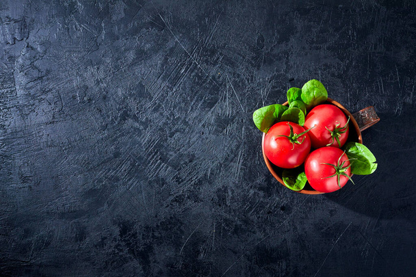 Ντομάτες φρέσκες με σπανάκι Φύλλα σε μαύρο πέτρινο φόντο. Χορτοφάγα λαχανικά διαίτης. Βότανα, κόκκινες ντομάτες, μαγειρική έννοια. - Φωτογραφία, εικόνα
