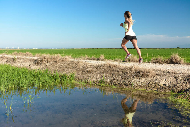 A young athlete girl in a yellow shirt running down a path between rice fields, with her reflection in the paddy water - Photo, Image