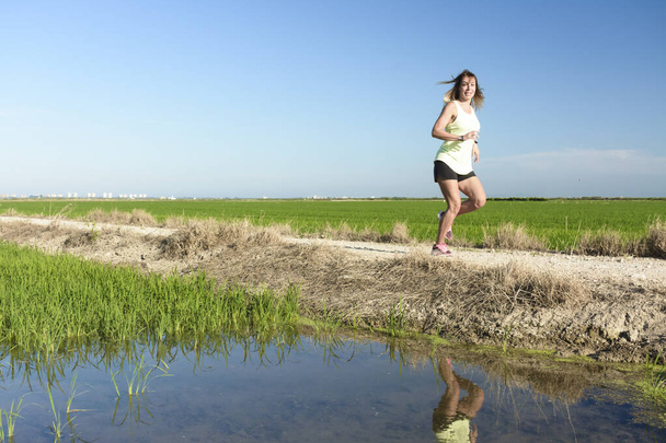 Sarı tişörtlü genç bir sporcu kız pirinç tarlaları arasında koşuyor, çeltik sularındaki yansımasıyla. - Fotoğraf, Görsel