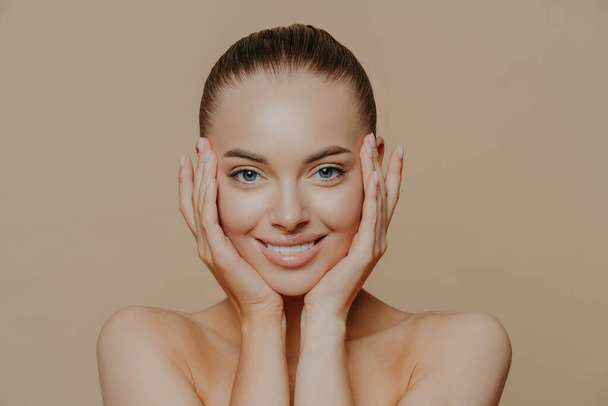 Skin care and beauty concept. Headshot of beautiful cheerful woman touches face gently, has perfect smile, healthy skin after cleaning or applying facial mask, isolated over beige background - Zdjęcie, obraz