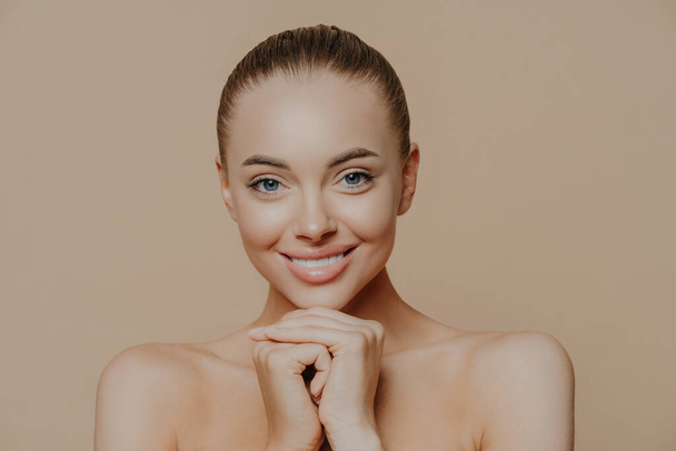 Close up shot of adult woman with fresh daily makeup, smiles toothily and keeps hands under chin, stands half naked indoor, feels refreshed after cosmetic procedures. Beauty and face care concept - Photo, Image