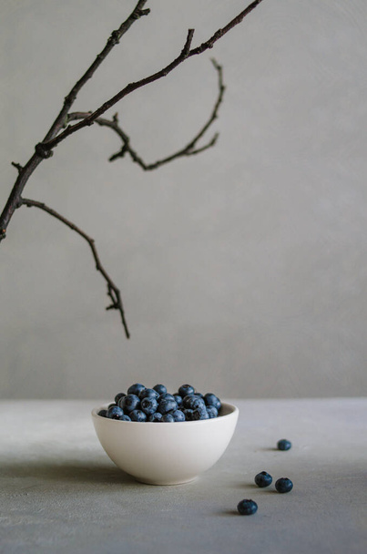 Bleuet frais mûr dans un bol blanc sur fond gris avec une branche noire. Concept de nourriture noire  - Photo, image