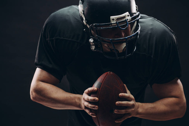 American football player with helmet and armour with a ball against black wall - Foto, Imagen