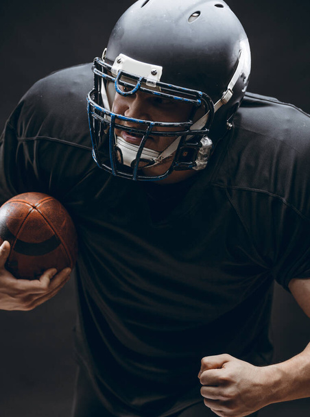 American football player with helmet and armour with a ball against black wall - Fotoğraf, Görsel