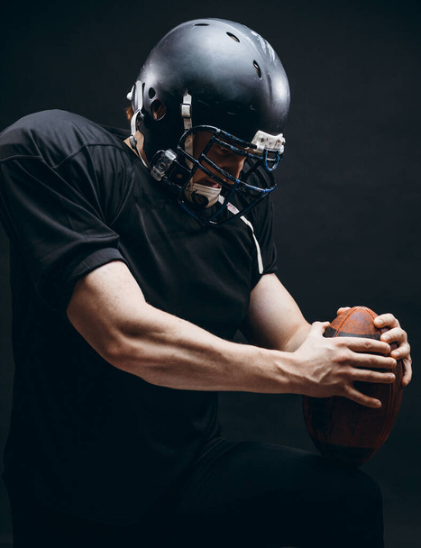 American football player with helmet and armour with a ball against black wall - Valokuva, kuva