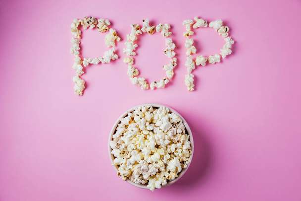 A pink bowl of popcorn on pink background, word POP made of corn, top view - Fotó, kép