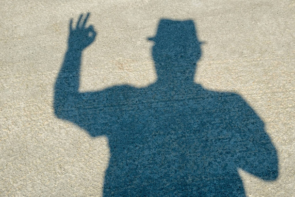 Schaduw van een man met een hoed op de betonnen vloer. Menselijke handen tonen symbolen Donker silhouet uit de schaduw. Tekenen: Oké, vijf, daar, overwinning. - Dag. Zonnig. Georgië. - Foto, afbeelding