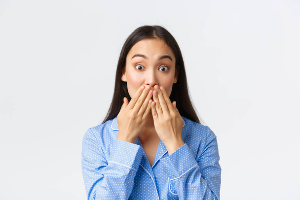 Close-up of shocked and astounded cute asian girl in blue pajama realise something, holding hands on mouth and pop eyes amazed at camera, hear gossips, white background - Foto, Bild