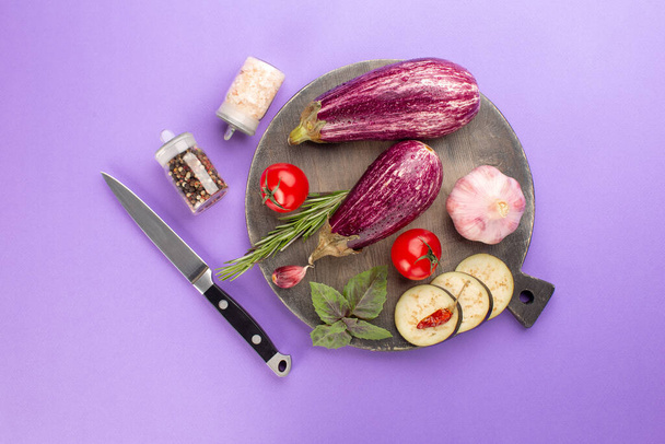 Summer vegetables on wooden board on purple background:ripe eggplants,tomatoes,fresh rosemary,purple basil leaves,garlic and hot pepper.Top view - Foto, Imagem