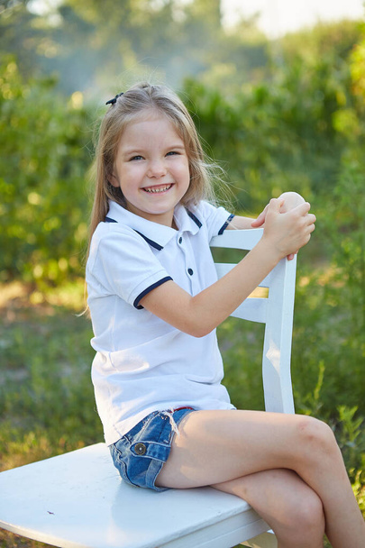 Küçük sevimli kız yaz bahçesinde beyaz bir sandalyede oturuyor. - Fotoğraf, Görsel