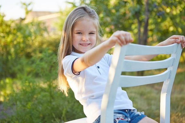 Маленькая очаровательная девочка сидит на белом стуле в летнем саду - Фото, изображение