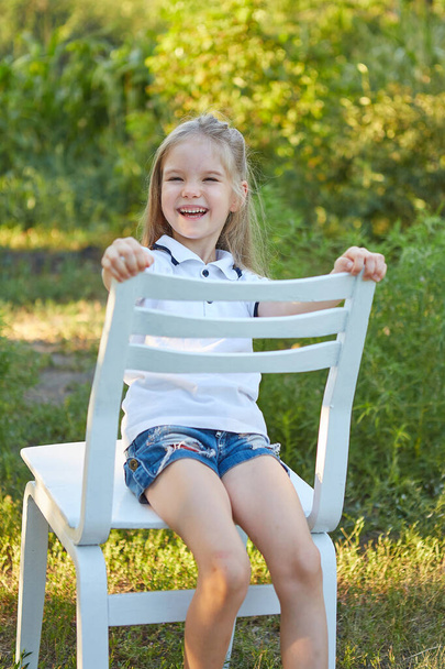 Kis bájos lány ül egy fehér széken a nyári kertben - Fotó, kép