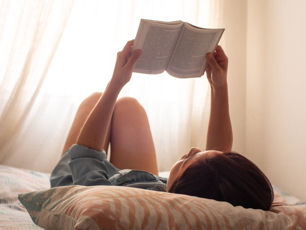 Молодая латиноамериканка читает книгу, лежащую на кровати, поддерживаемую подушкой из листьев. - Фото, изображение