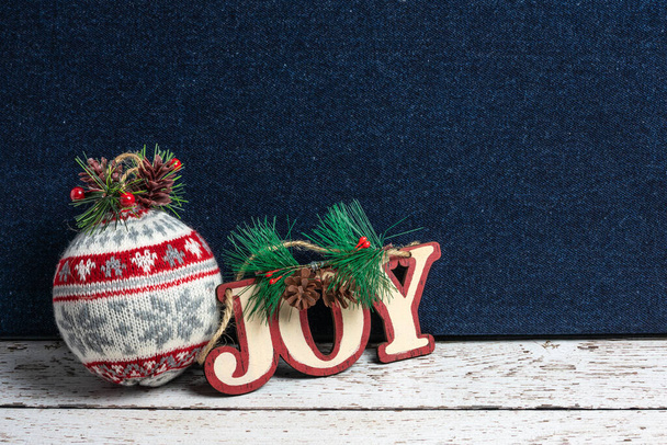 Saludo navideño del país con un adorno de bola de punto y un recorte de madera "Joy" con verdes y piñas sobre una mesa de madera pintada de blanco rústico y un fondo de denim azul
 - Foto, imagen