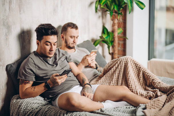 Feliz pareja gay acostados juntos en la cama y el uso de teléfono inteligente, mirando las fotos
 - Foto, imagen