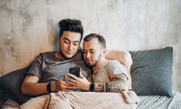 Happy gay couple lying together in bed and using smartphone, looking at photos - Фото, изображение