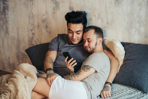 Happy gay couple lying together in bed and using smartphone, looking at photos - Foto, Imagem