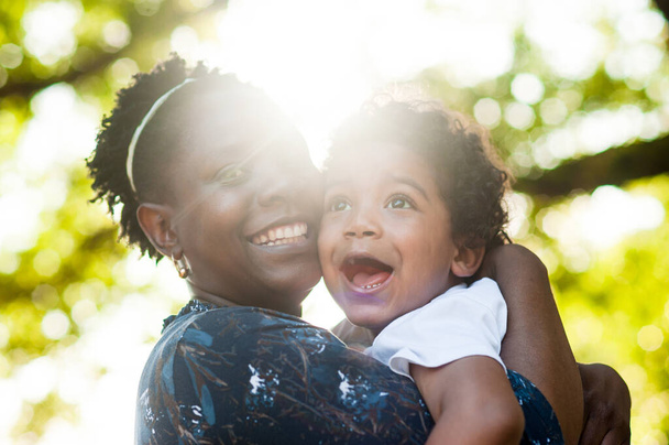 Afrikkalainen äiti hymyilee kameralle pitäen pikkupoikaansa sylissään. Auringonvalo valaisee paikan. Luonnollinen liekki - Valokuva, kuva