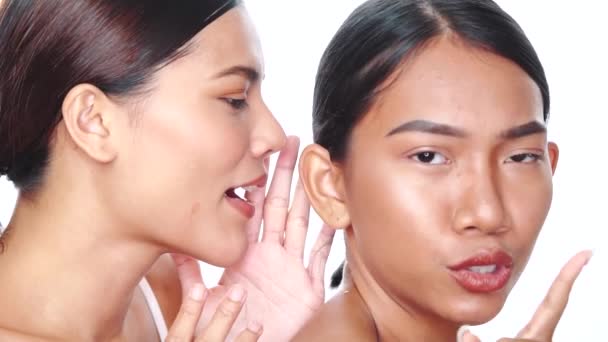 Close-up gezichten van twee mooie Aziatische vrouwen roddelen over witte achtergrond. Vriend vertelt een geheim aan vriendin fluisterend in het oor - Video