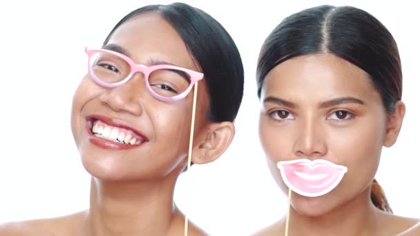 Portret van twee jonge lachende Aziatische vrouwen die poseren met rekwisieten bril en lippen over een witte achtergrond. Feest, fotohokje, vriendschap, leuk tijdconcept - Video