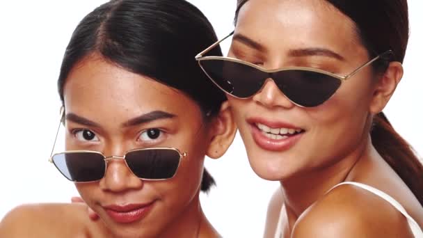 Портрет двух молодых улыбающихся азиатских женщин, улыбающихся, позирующих в солнцезащитных очках на белом фоне - видео в замедленной съемке - Кадры, видео