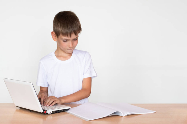 A cute boy sits at a table, prints on a laptop, looks into a notebook or book on a white background. Concept Back to school. Education concept. Copy space - Photo, Image