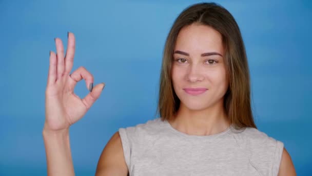 Beautiful woman in gray t-shirt smiles, raises hand, shows ok on blue background - Footage, Video